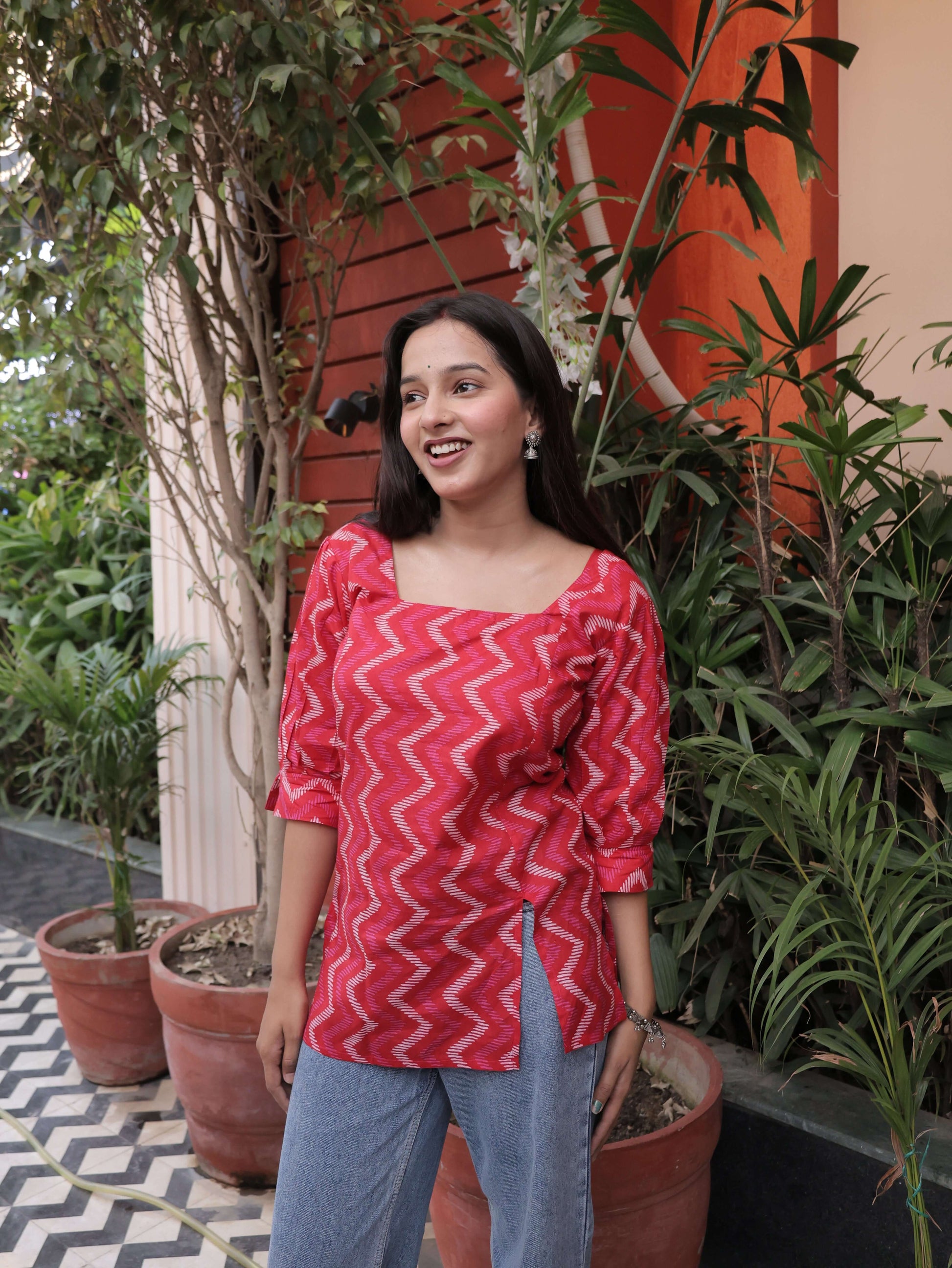 Woman in a red chevron-patterned dress standing outdoors with plants and a patterned floor.